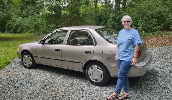 Woman stands next to a gold Toyota Corolla on a gravel driveway, with trees in the background.