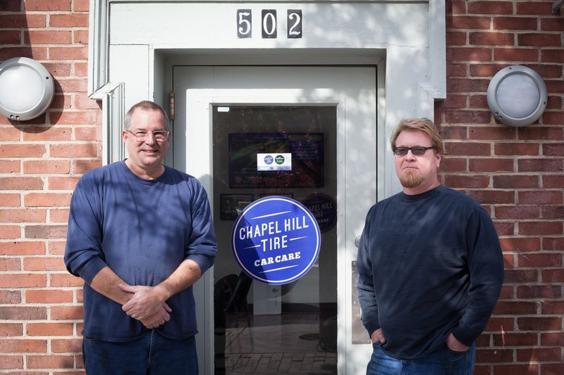 Two men stand in front of a Chapel Hill Tire door. The building is brick, with the number 502 above the door.