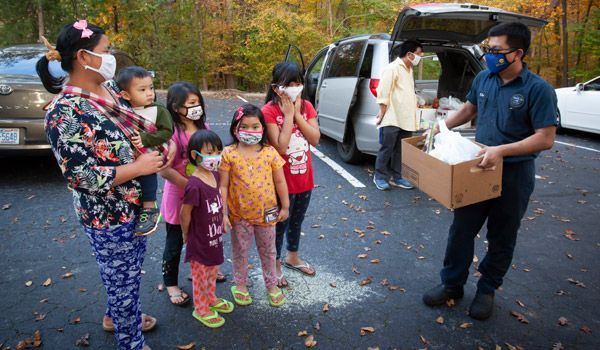 Man gives food box to family, all wearing masks, near parked van in outdoor setting.