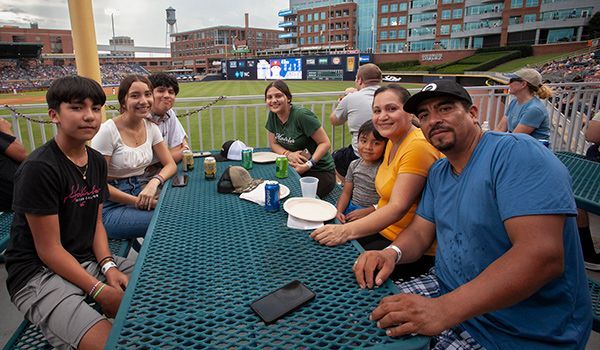 Family at a baseball game, sitting at a picnic table, smiling, looking at the camera.