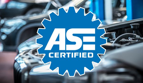 ASE Certified logo overlaid on a blurry auto repair shop scene. Blue cogwheel with white text.