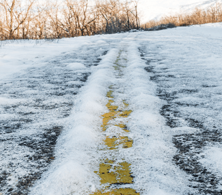 Snow-covered road with visible yellow lane marking, trees in background, sunny day.