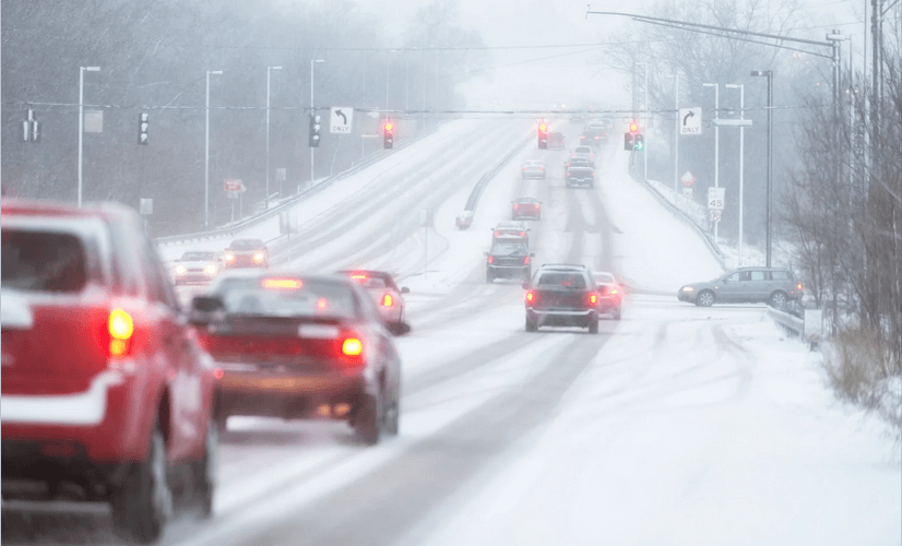 Snow-covered road with vehicles driving through the falling snow; traffic lights are visible.