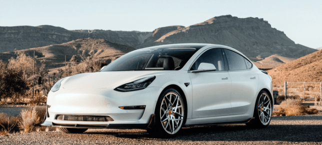 A white tesla model 3 is parked on the side of a road with mountains in the background.