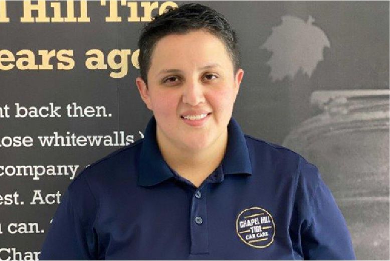 Woman in a blue polo shirt smiles in front of a sign for a tire company.