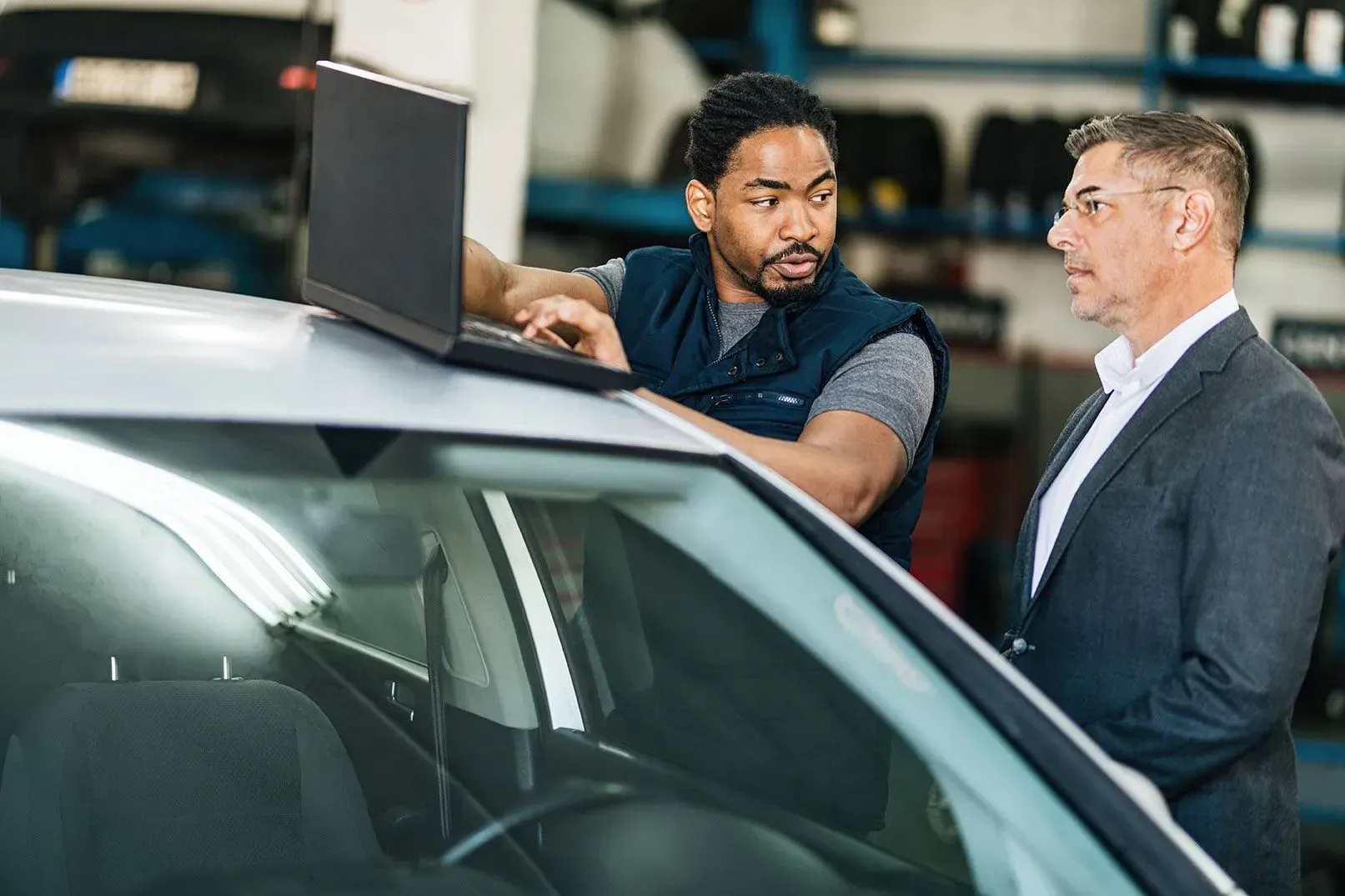 A technician with a laptop on a car roof discusses vehicle diagnostics with a customer in a repair shop.
