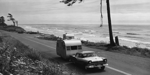 A black and white photo of a car pulling a trailer down a road.