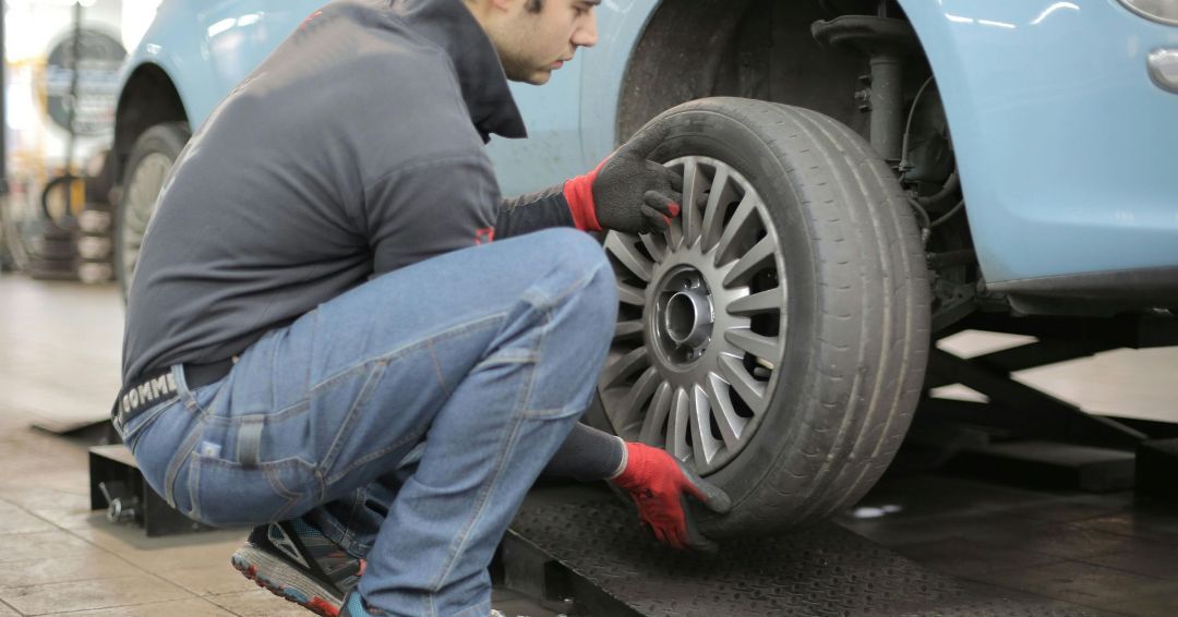 Mechanic in gray shirt and jeans changes a car tire on a ramp; light blue car.