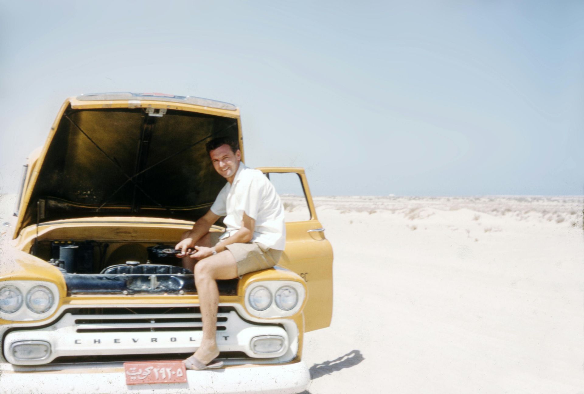 Man sitting on a yellow truck with hood open in a desert landscape.