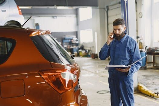 Mechanic in blue overalls on phone, holding clipboard, near a reddish-orange car inside a garage.