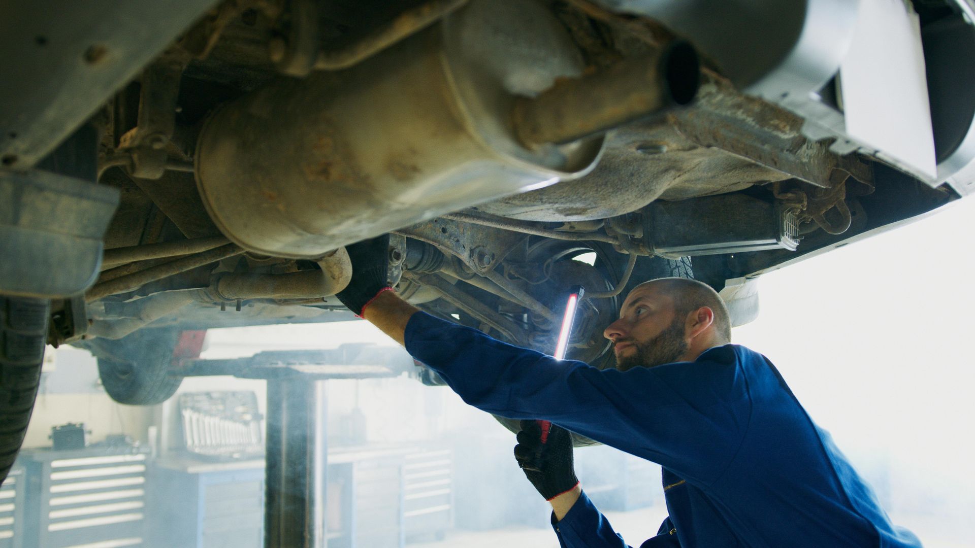 Mechanic in blue coveralls repairing the underside of a vehicle on a lift in a workshop