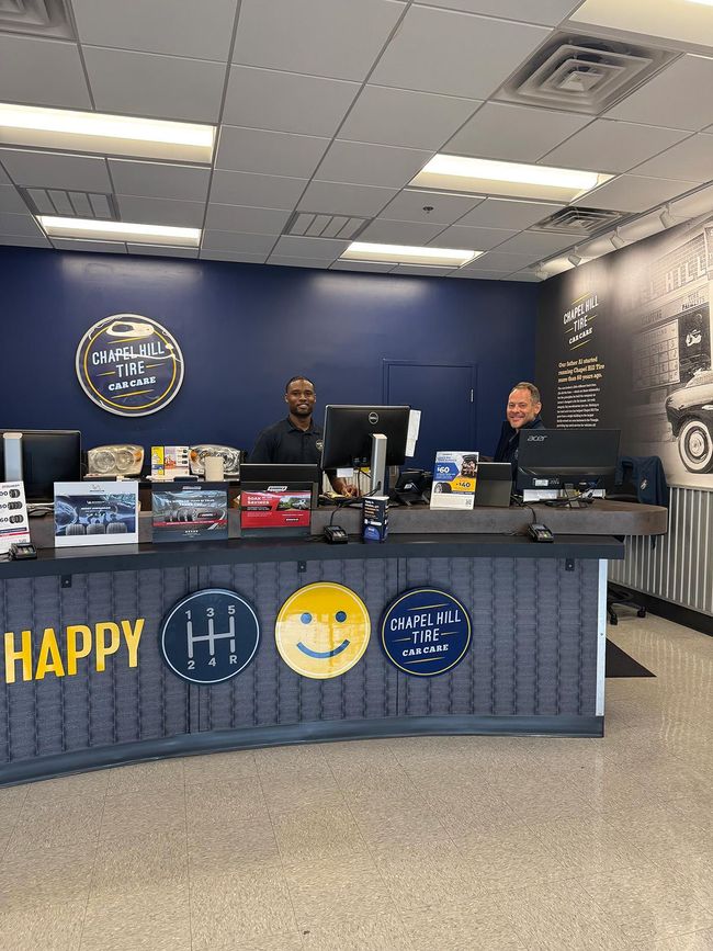 Two men at a car repair shop front desk. Dark blue and yellow decor, smiley face emblem, computer monitors.