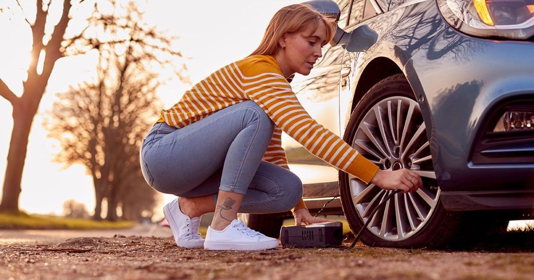 Woman inflating a tire on a blue car. She kneels on a roadside with a pump. Sunset light.
