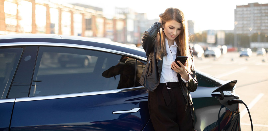 Woman standing beside a dark blue car, looking at her phone while charging it outdoors.