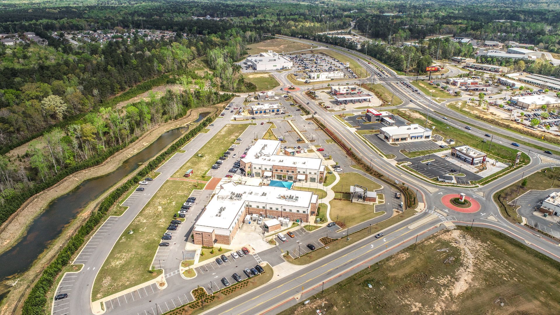 Aerial view of a suburban shopping complex with parking lots, roads, and surrounding trees.
