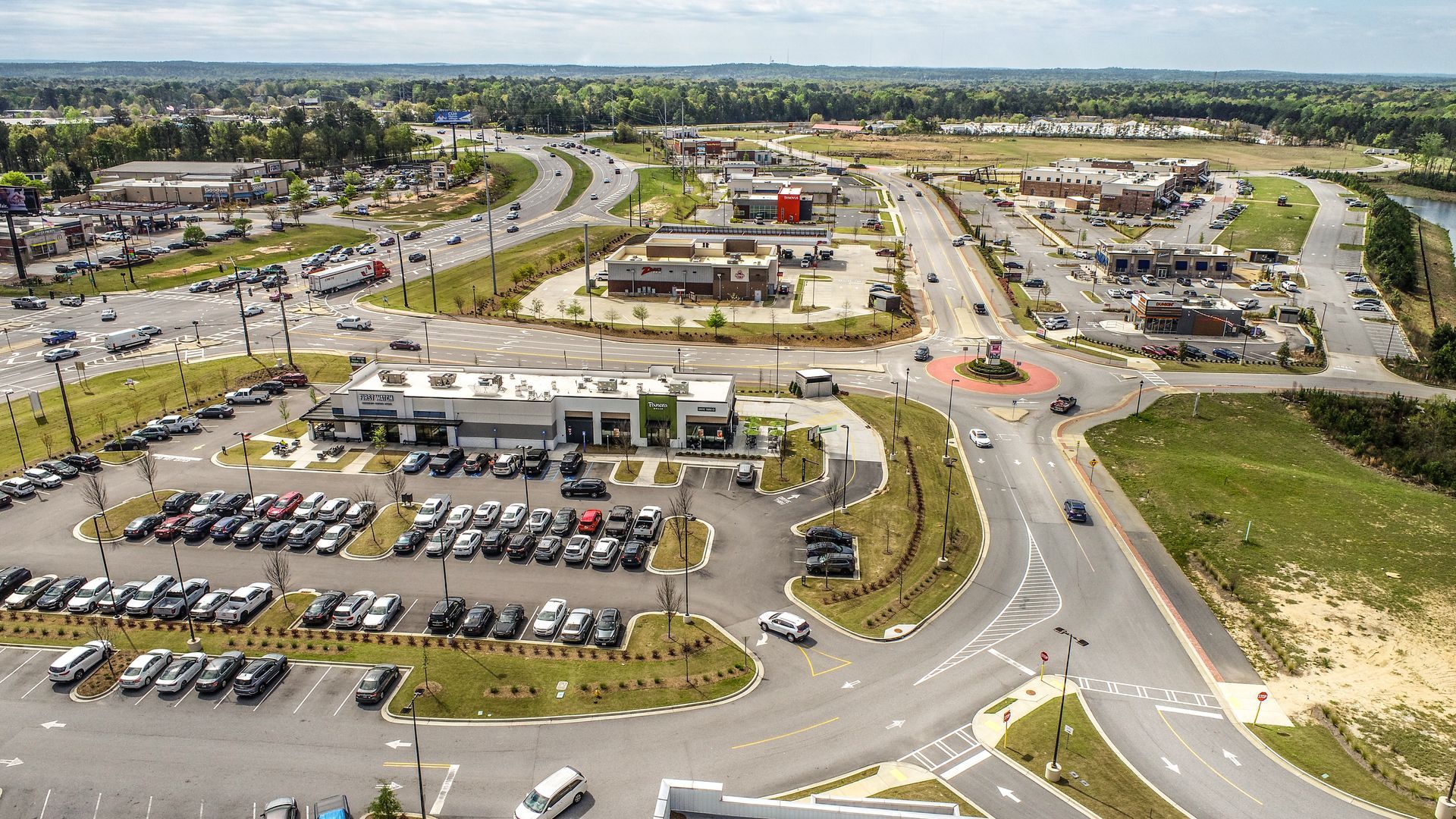 Aerial view of a large parking lot, road intersection, and commercial buildings in a suburban area.