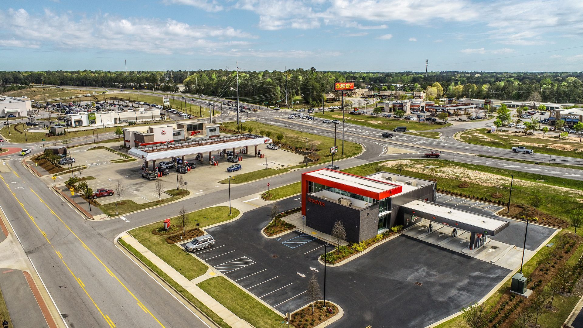 Aerial view of a gas station, parking lot, and nearby road intersection on a sunny day.