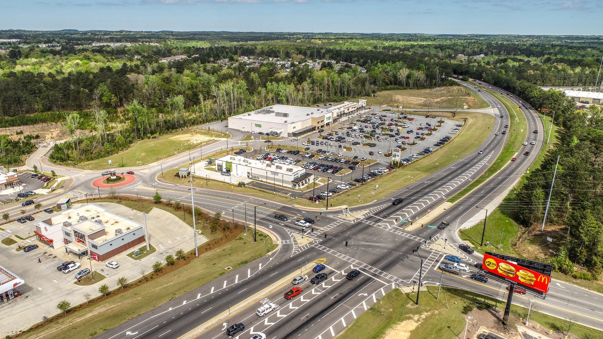 Aerial view of a busy highway interchange beside a large parking lot and commercial buildings, surrounded by greenery.