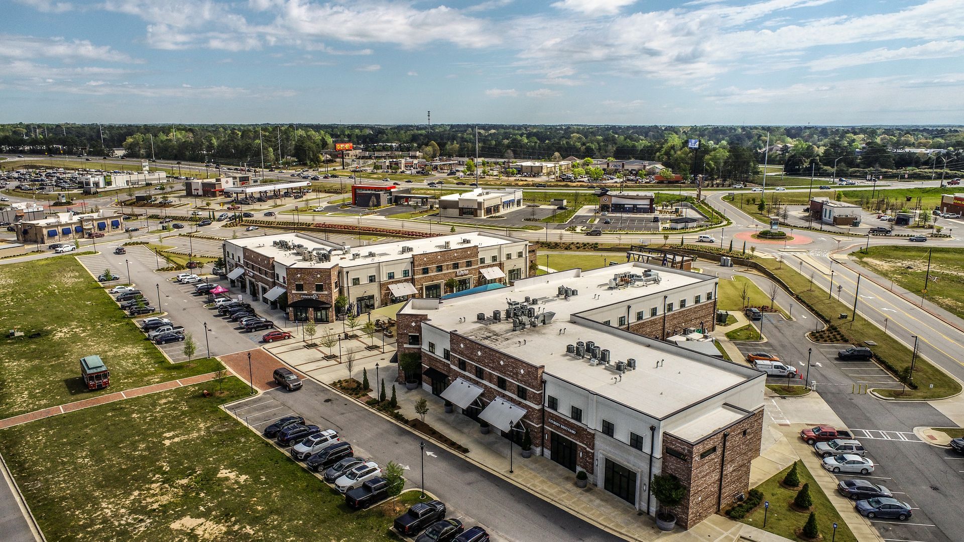 Aerial view of a shopping center with parking lots, storefronts, and surrounding roads under a clear sky