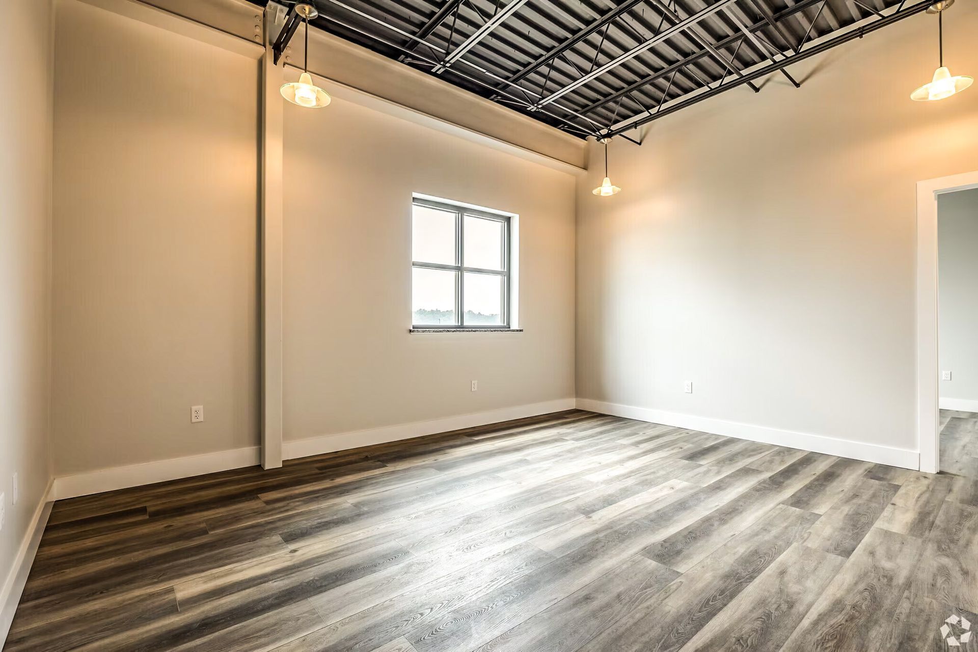 Empty modern room with white walls, exposed ceiling, and wood-look flooring, lit by warm lights.