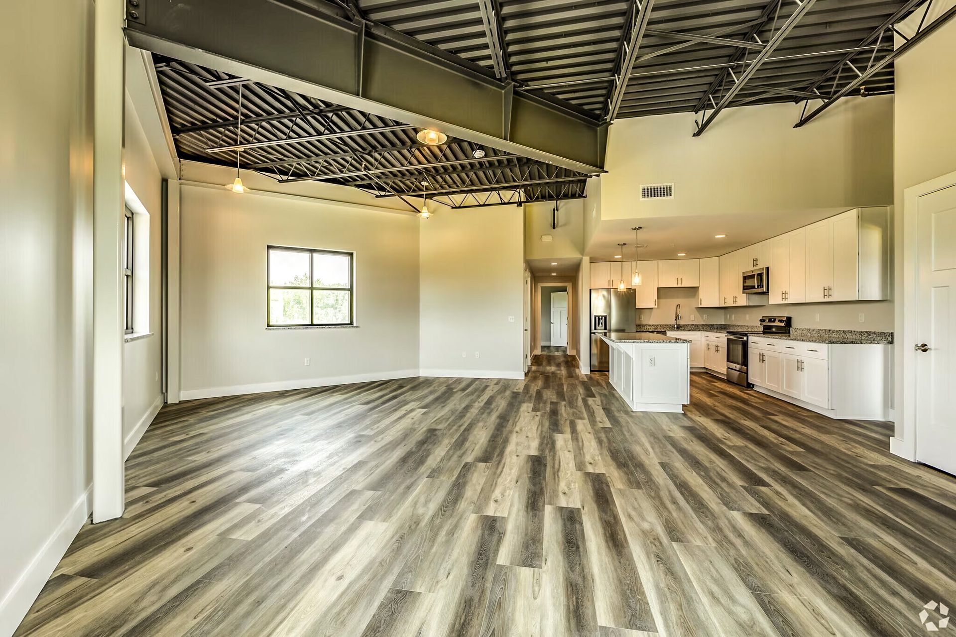 Spacious empty loft with exposed ceiling beams, wood-look flooring, and a small kitchen on the right.