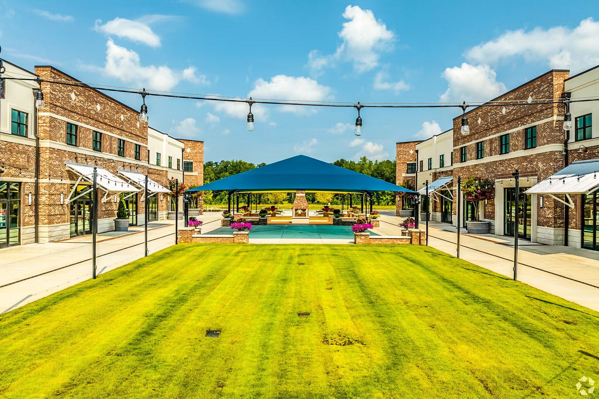Wide green lawn between brick storefronts, with string lights and a blue canopy at the far end.
