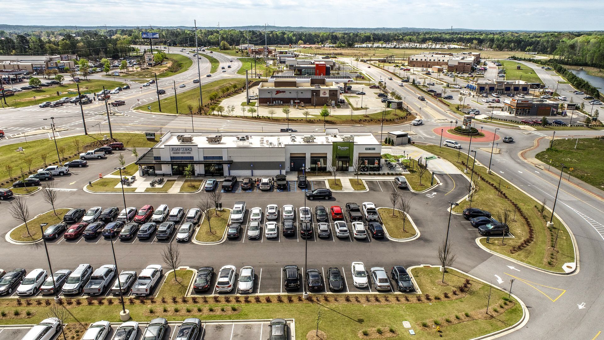 Aerial view of a shopping center parking lot with many cars, roads, and surrounding businesses.