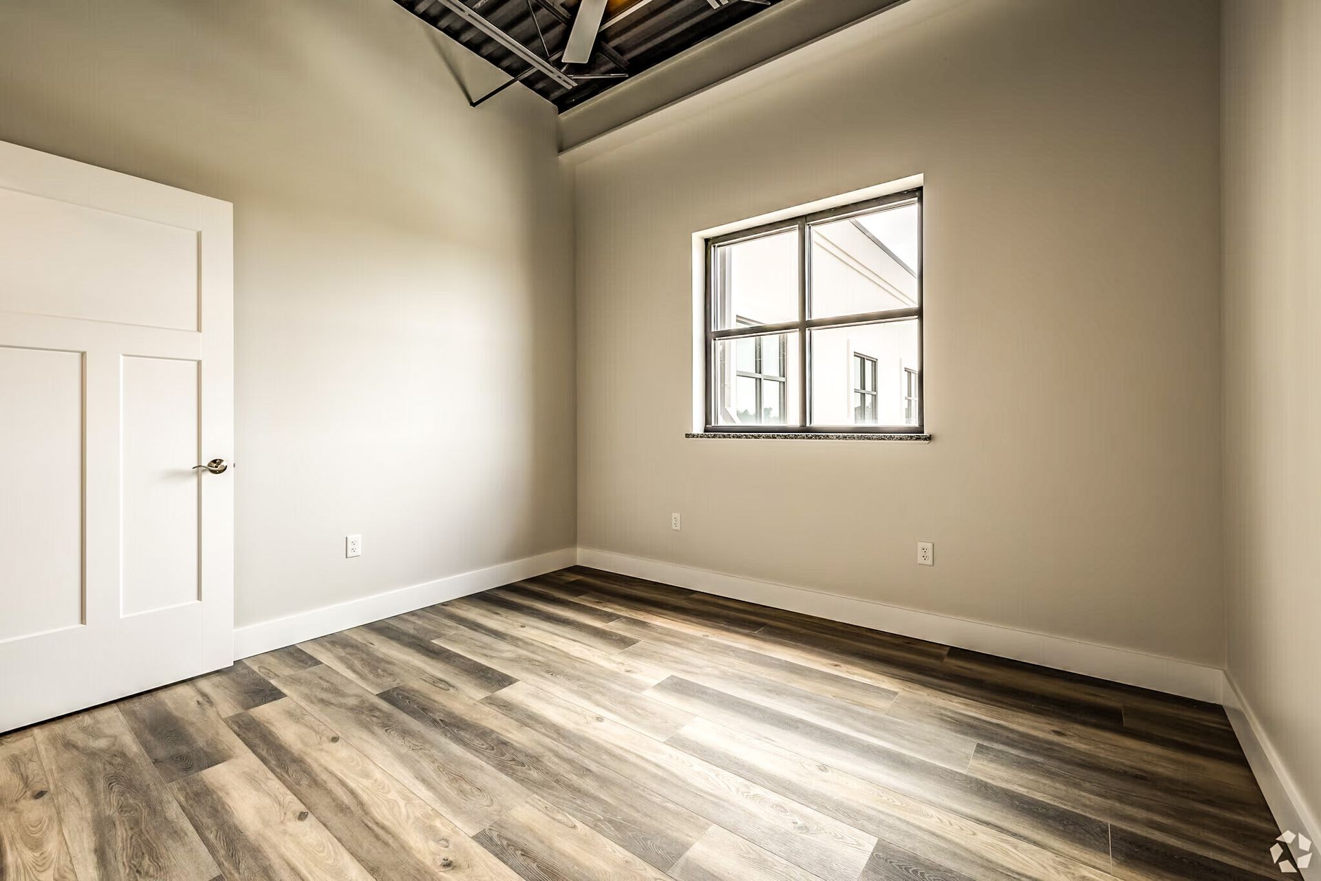 Empty beige room with wood flooring, white door, and a small window near the ceiling