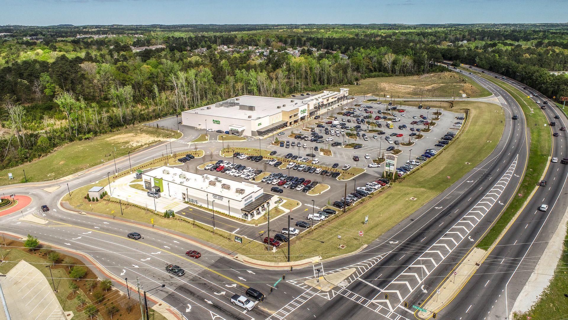 Aerial view of a large shopping center, parking lot, and highway surrounded by trees.