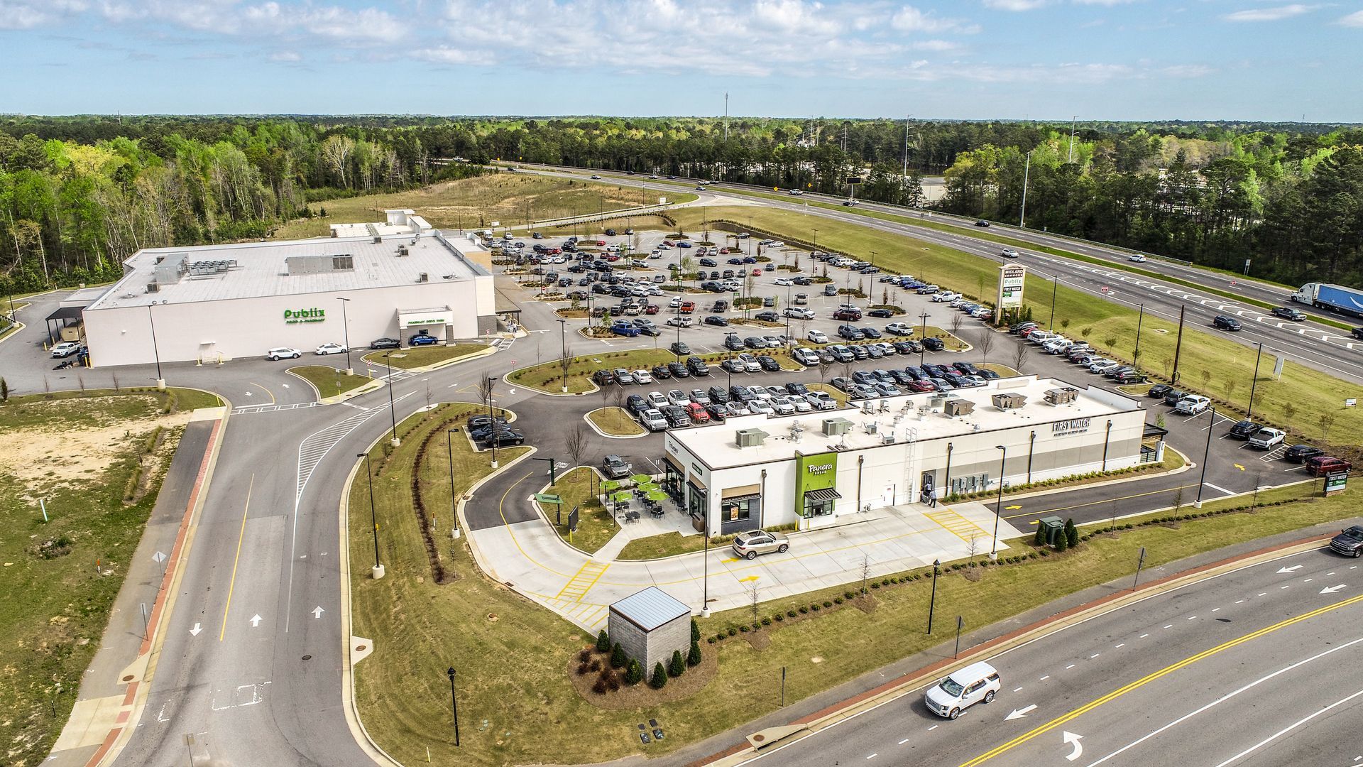 Aerial view of a large commercial building and parking lot beside a highway, surrounded by trees.