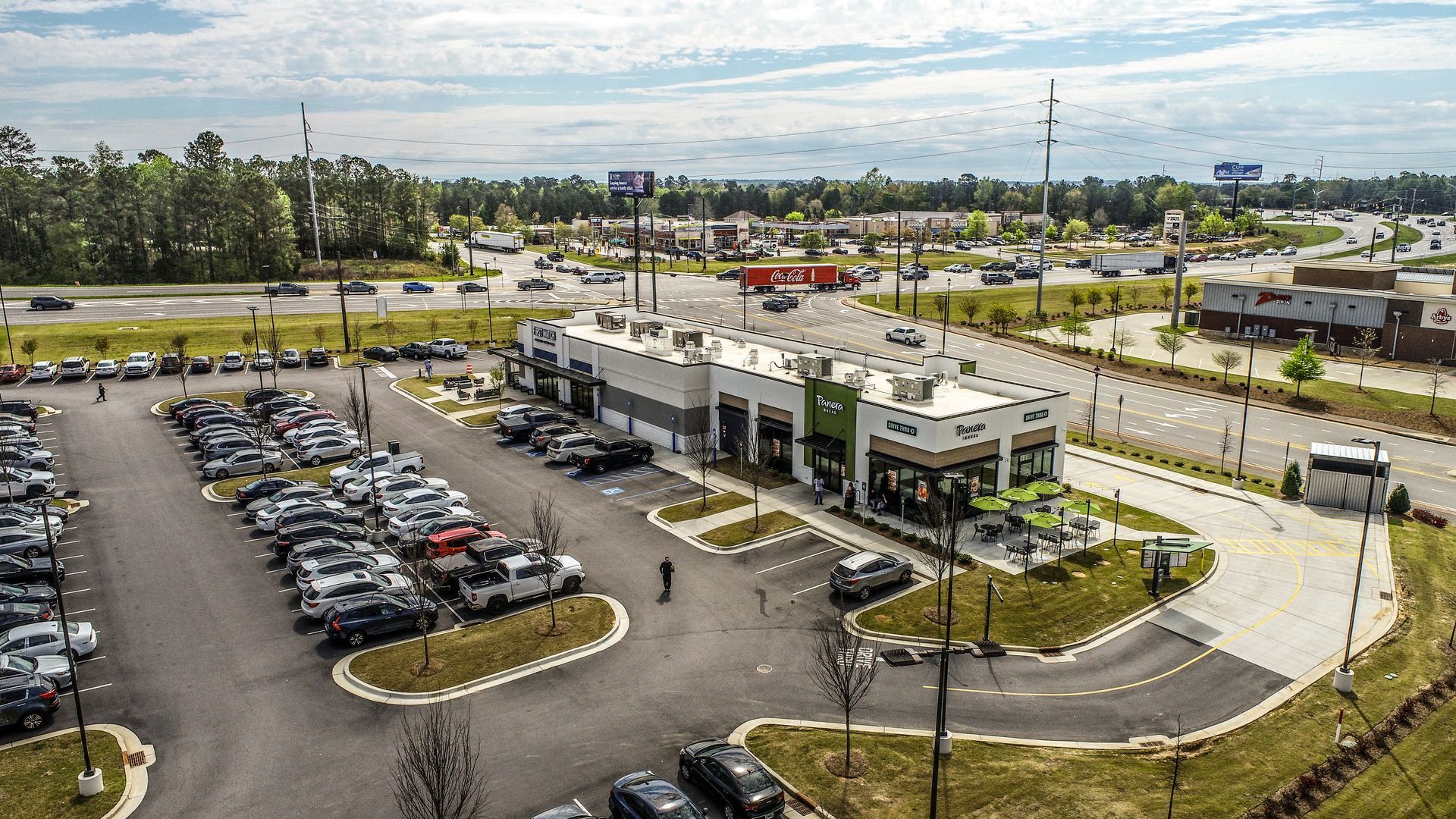 Aerial view of a shopping plaza with parking lots, storefronts, cars, and a cloudy sky.