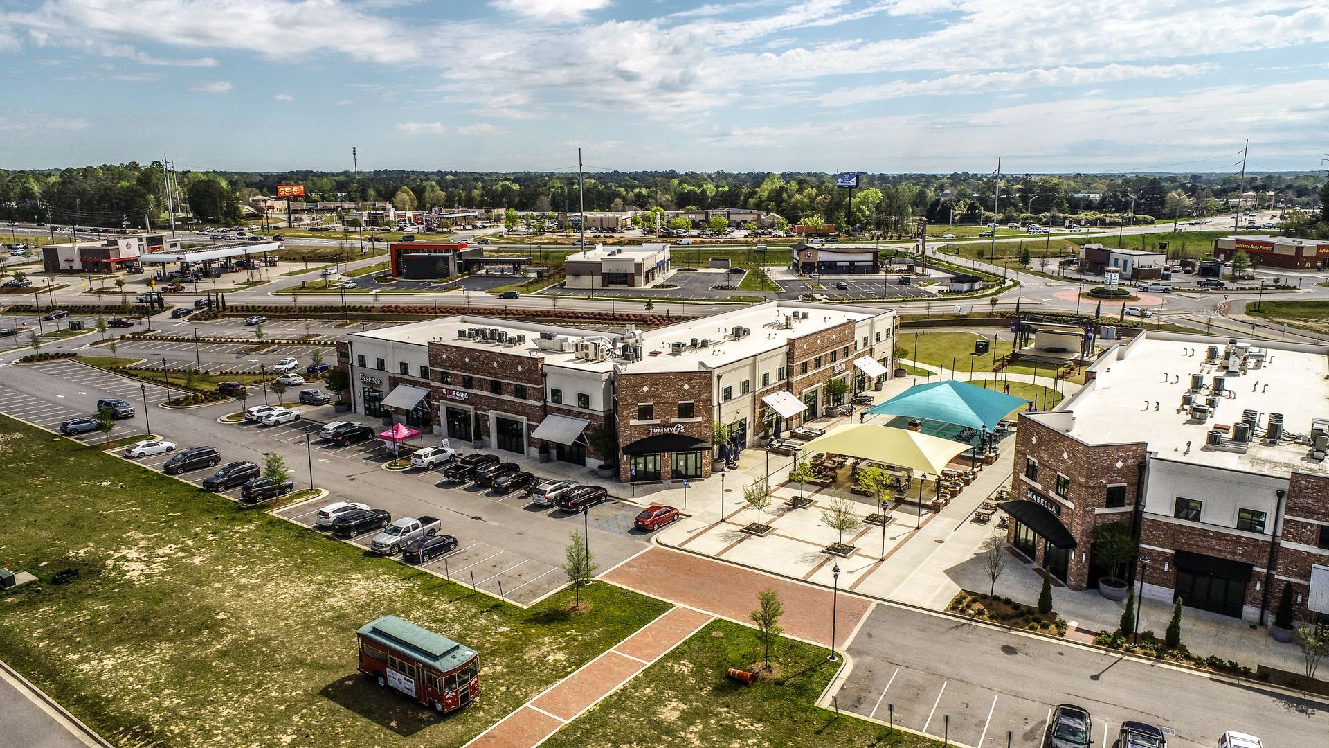Aerial view of a busy shopping center with parking lots, storefronts, and a partly cloudy sky