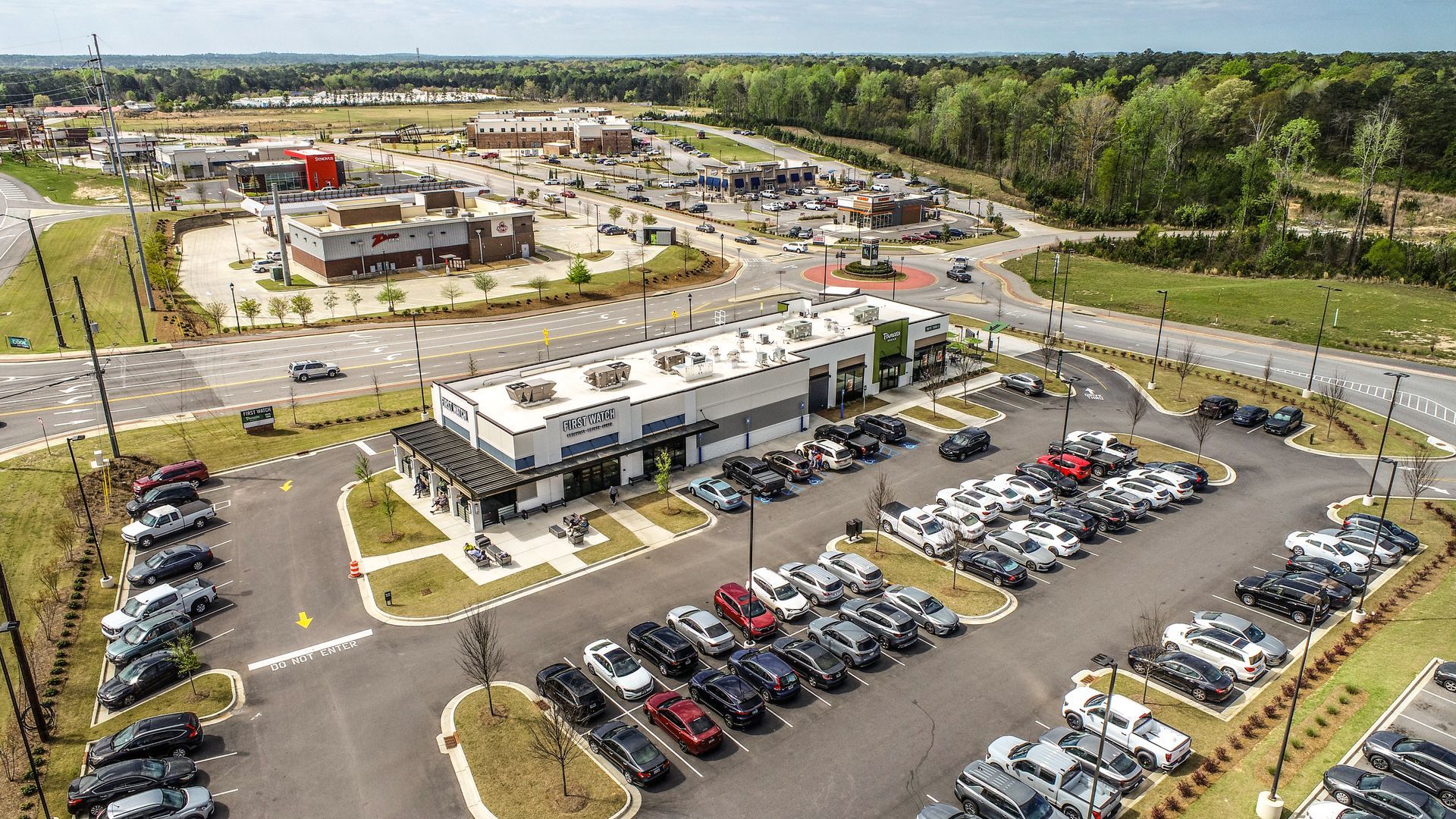 Aerial view of a large commercial building and parking lot with many parked cars, surrounded by roads and greenery.