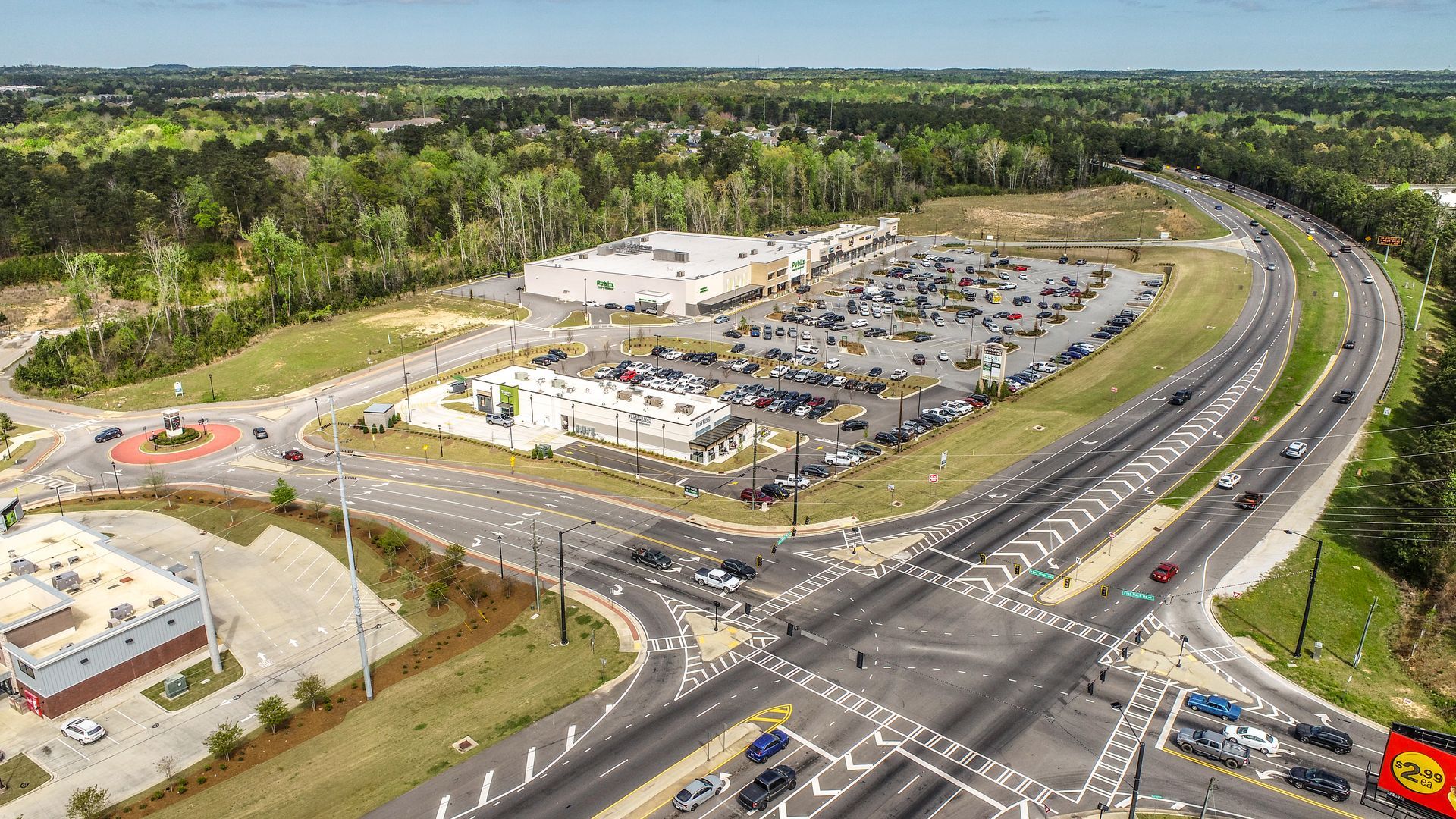 Aerial view of a shopping center and parking lot beside a multi-lane highway and green wooded area