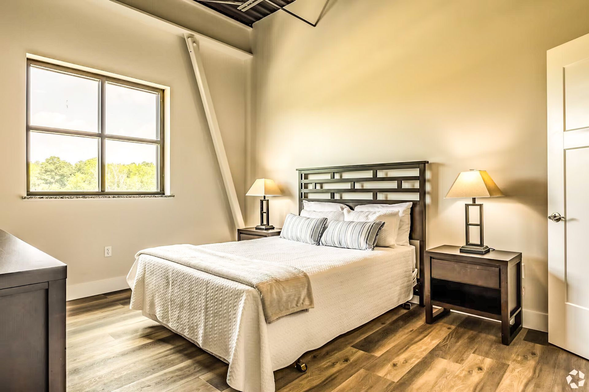 Bright bedroom with a made bed, bedside lamps, window, and wood floors