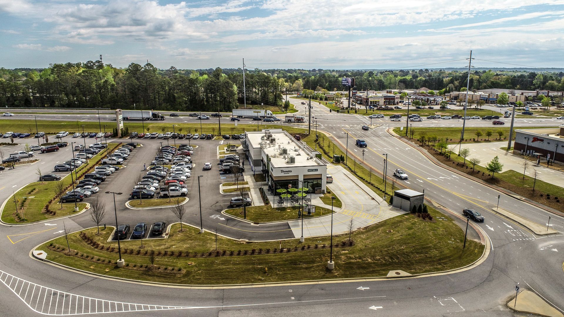 Aerial view of a parking lot, landscaped roundabout, and low buildings beside a road and green fields.