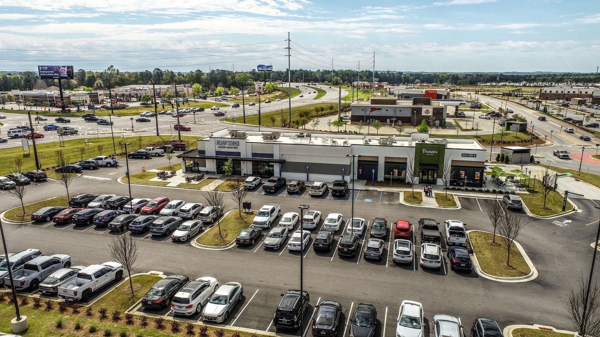 Aerial view of a busy shopping plaza with parking lots, cars, and surrounding roads under a partly cloudy sky