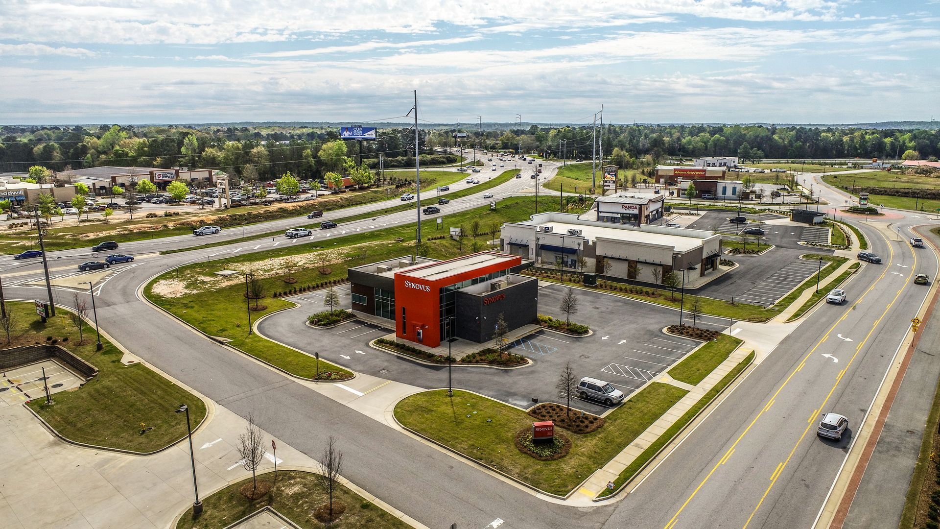 Aerial view of a roadside commercial area with a red-roofed building, parking lot, and curving intersection.