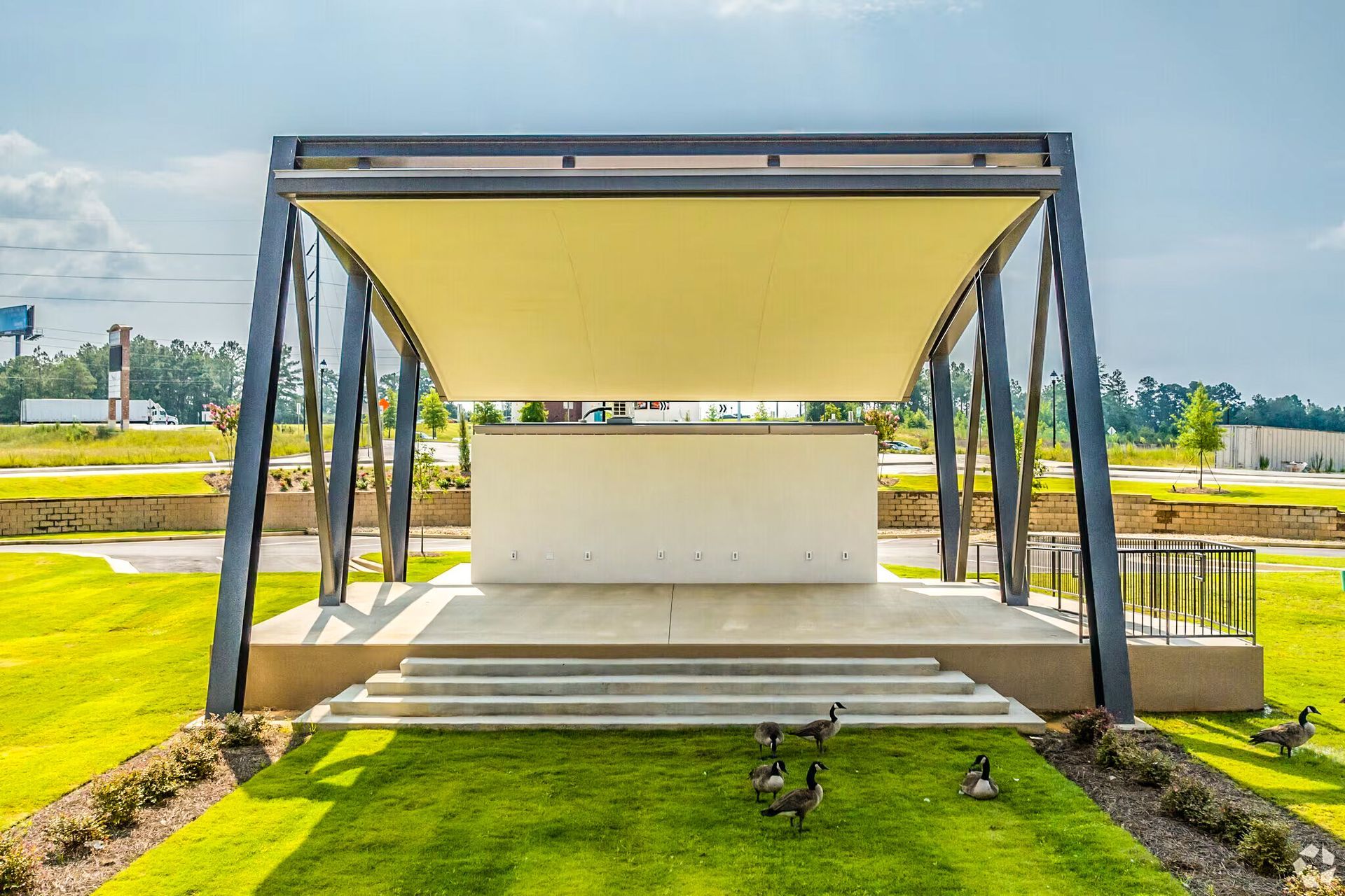 Outdoor stage pavilion with a yellow canopy, steps, and green lawn in a park.