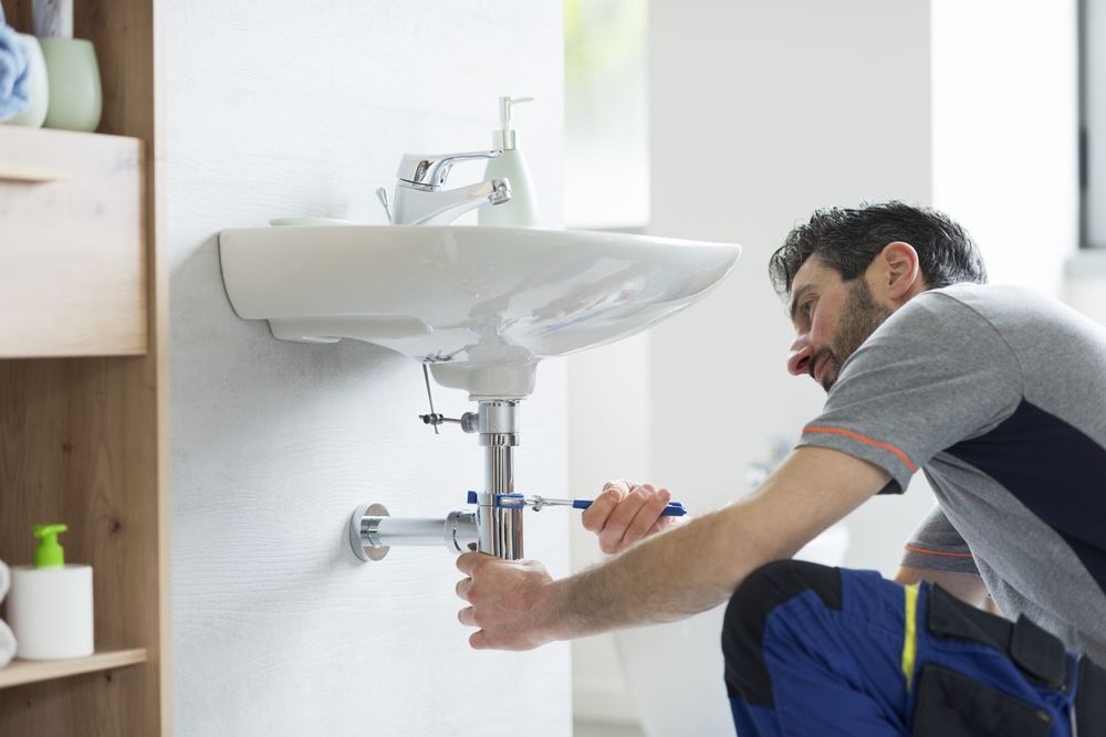 A plumber in work clothes kneeling in a bathroom, using a wrench to tighten the pipe underneath a wall-mounted sink.