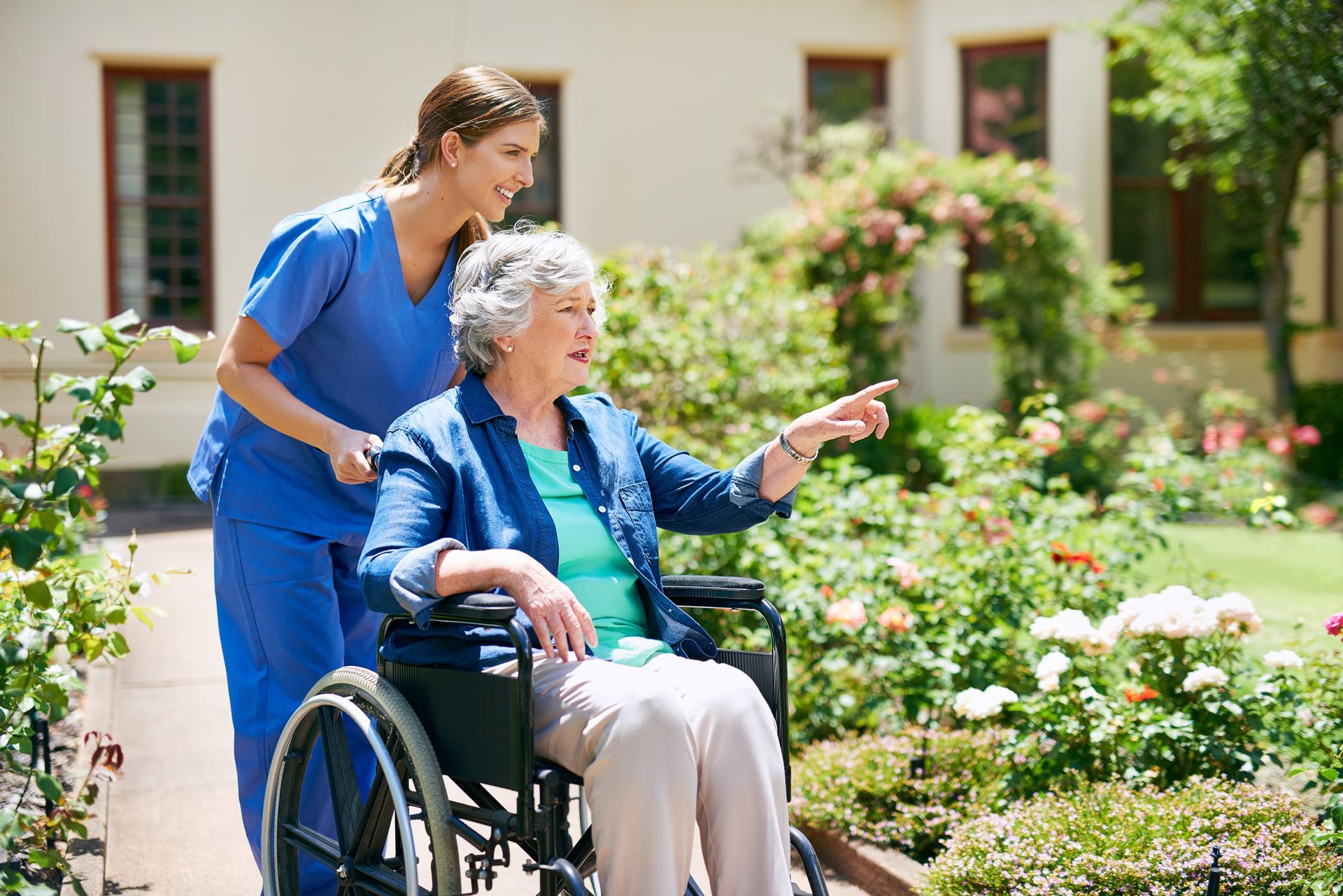 A caregiver in blue scrubs pushes an individual in a wheelchair through a sunny outdoor garden as they point ahead.