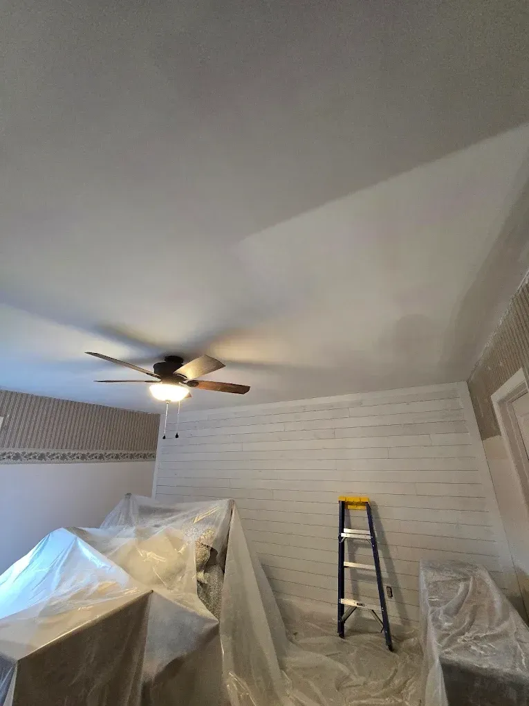 Room under renovation with plastic-covered furniture, ladder, ceiling fan, and freshly painted white wall