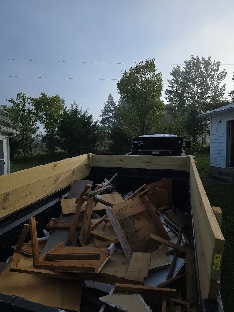 Truck bed filled with lumber and debris in a driveway, with trees and houses in the background