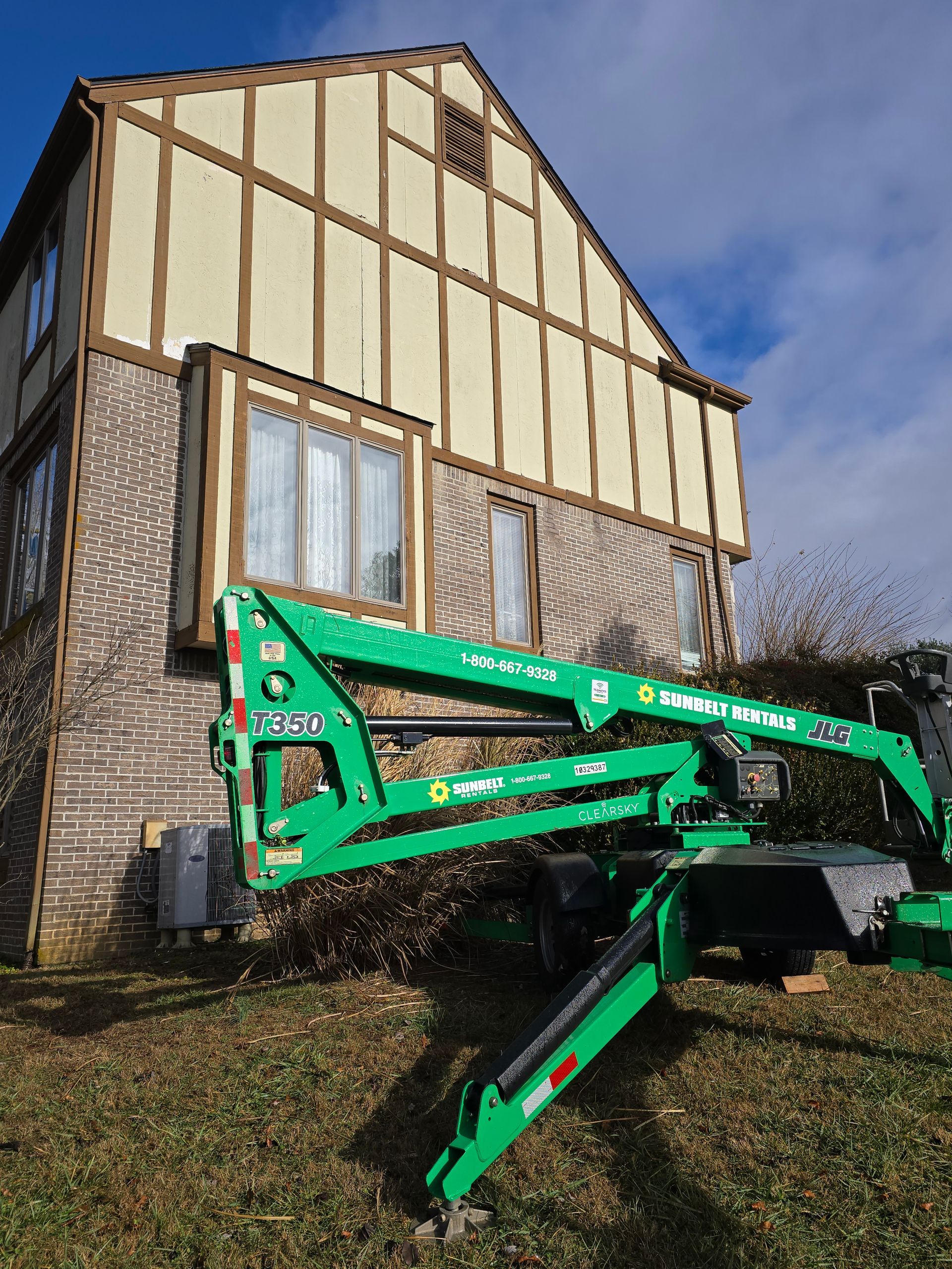 Green excavator parked in front of a small house with tan siding and a blue sky.