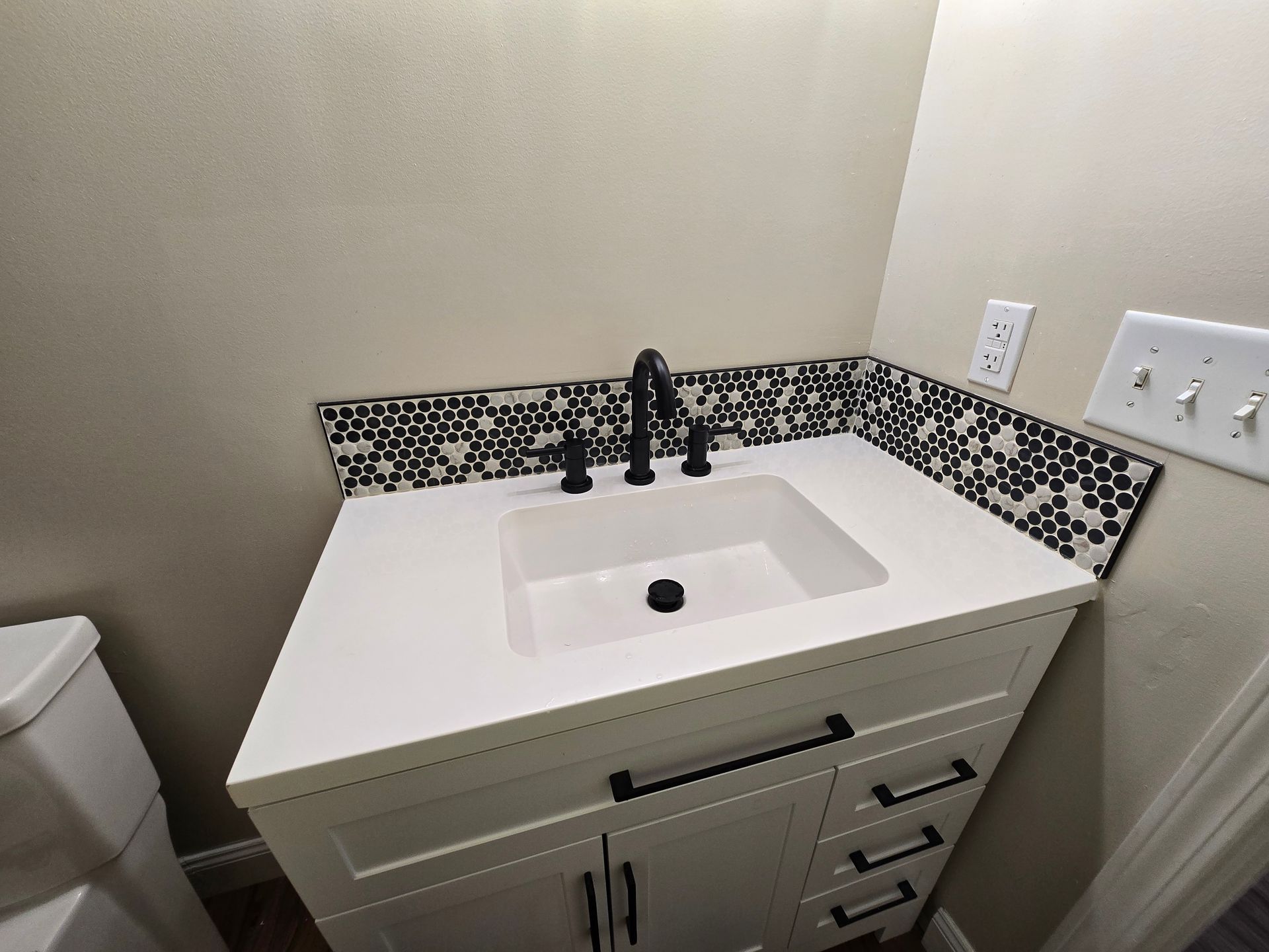 Bathroom vanity with white sink, black faucet, and mosaic tile backsplash beside a toilet