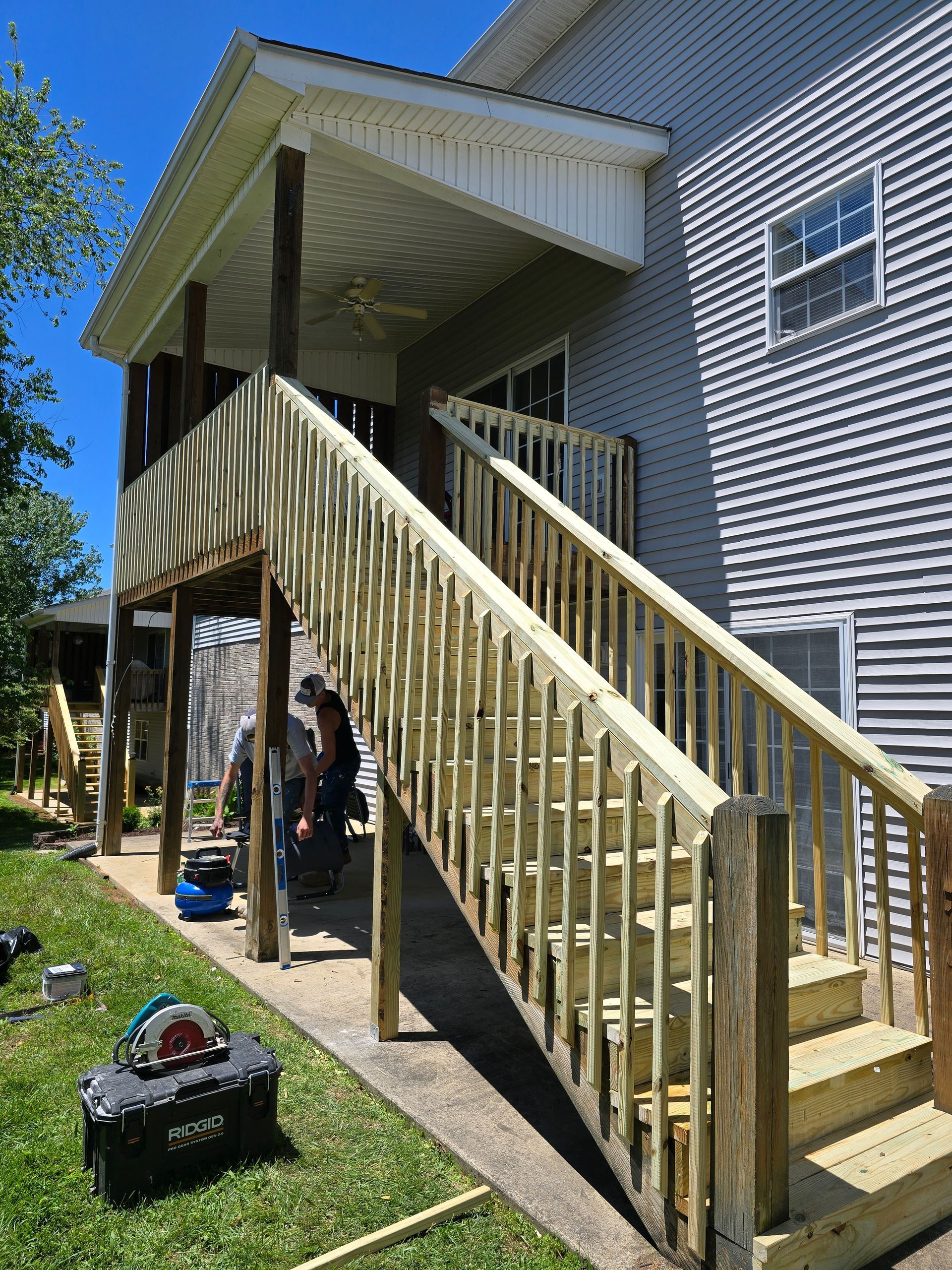 Wooden stairway and covered porch on a gray house, with people standing underneath on a sunny day