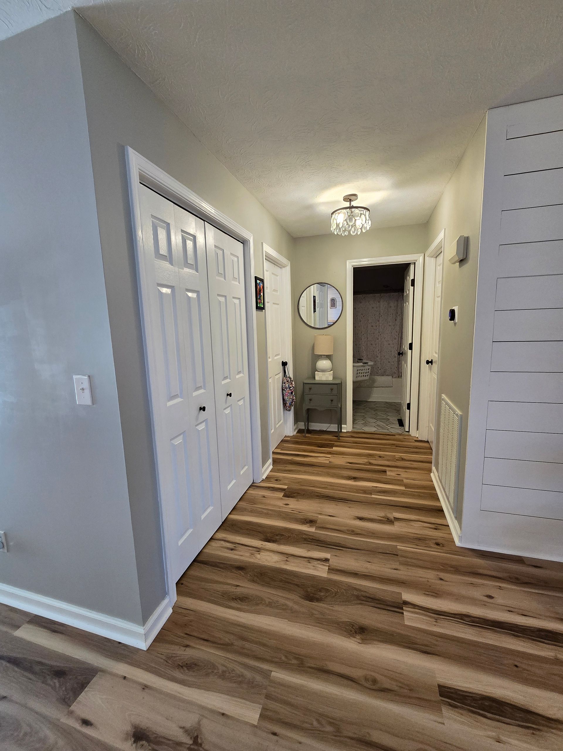 Hallway with wood flooring, white doors, mirror, and a small chair at the end.