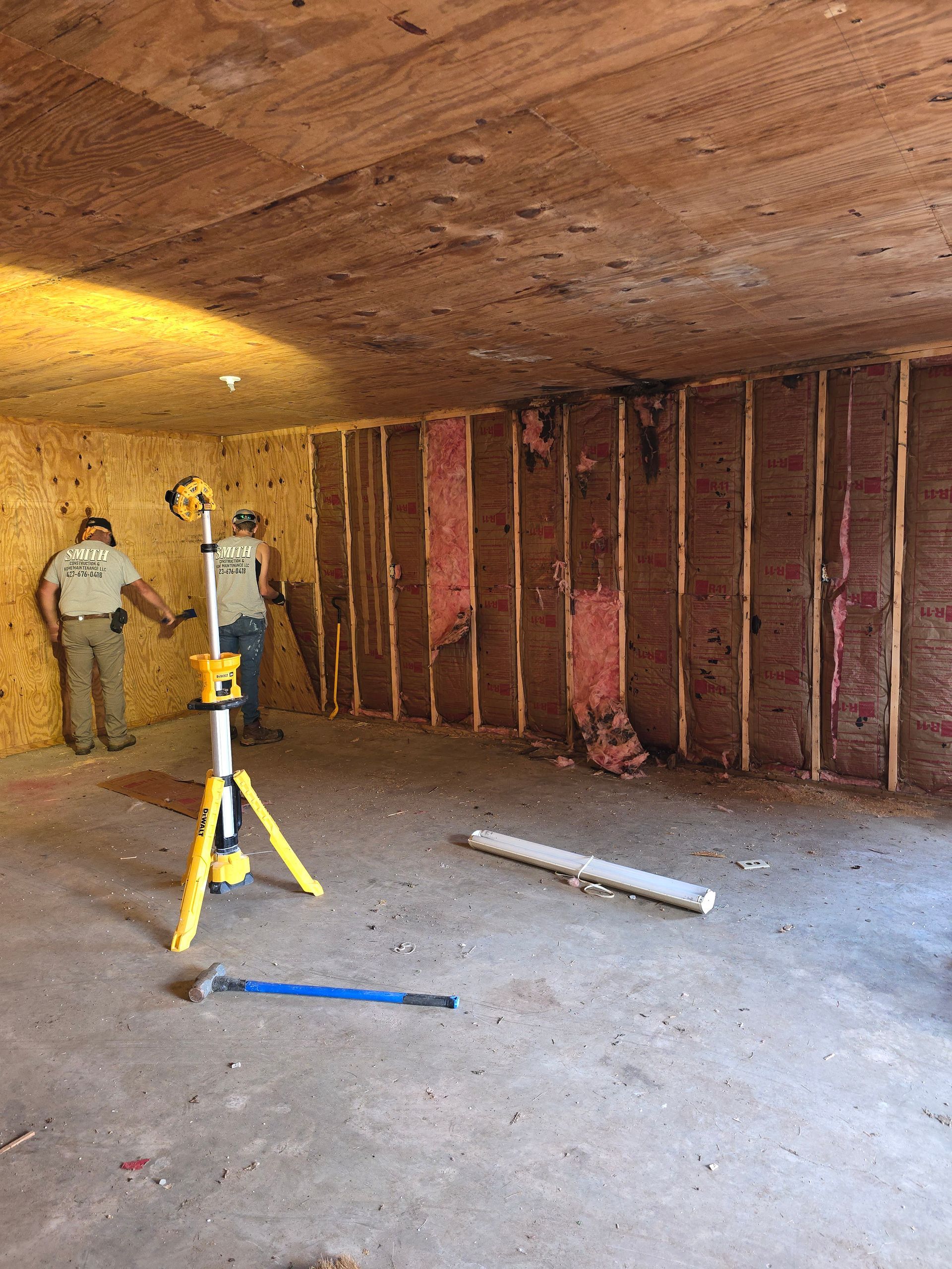 Two workers in a bare room with exposed wall studs and a yellow tripod laser level on the floor
