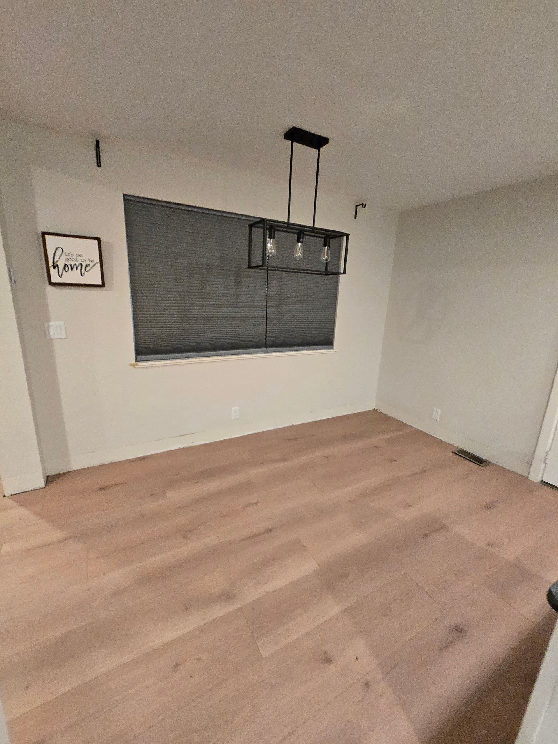 Empty room with light wood floor, white walls, dark window blinds, and a black pendant light fixture