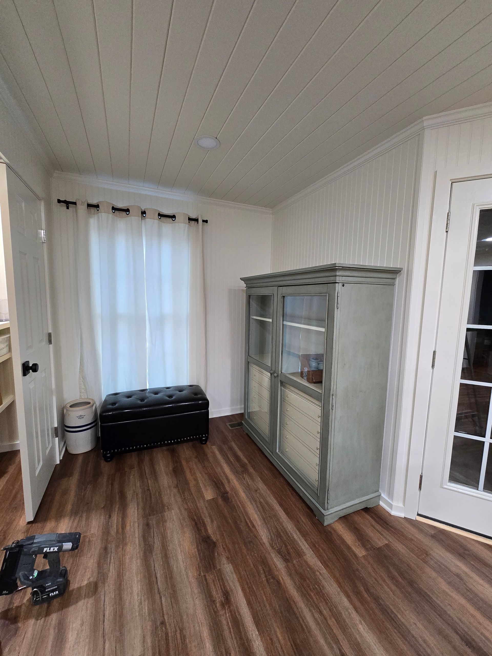 Small room with wood floor, sheer-curtained window, black bench, and gray display cabinet by a white door